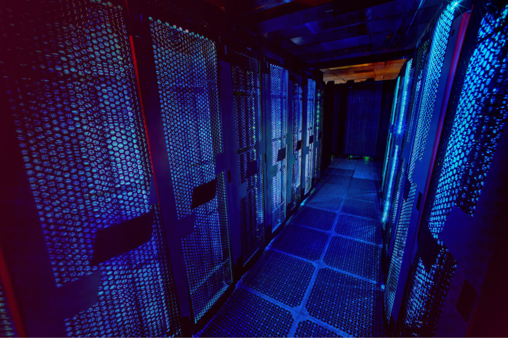 Serverrom med blå belysning og moderne teknologi Blått og lilla lys belyser en smal gang med serverkabinetter, hvor meshdører gir innsyn til teknologisk utstyr.
