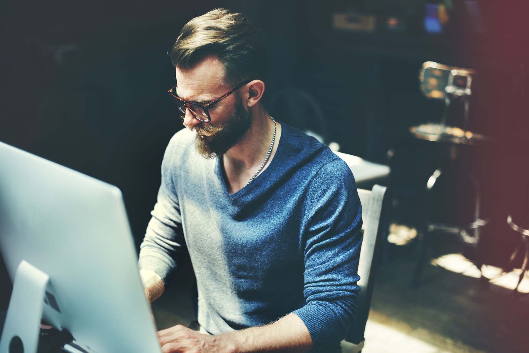 Tittel: Profesjonell webdesigner i arbeid ved datamaskin Beskrivelse: En mann arbeider med webdesign på en modern datamaskin. Mann med skjegg og briller sitter ved en datamaskin, iført en blå genser, i et stilrent arbeidsmiljø med svake lysforhold.