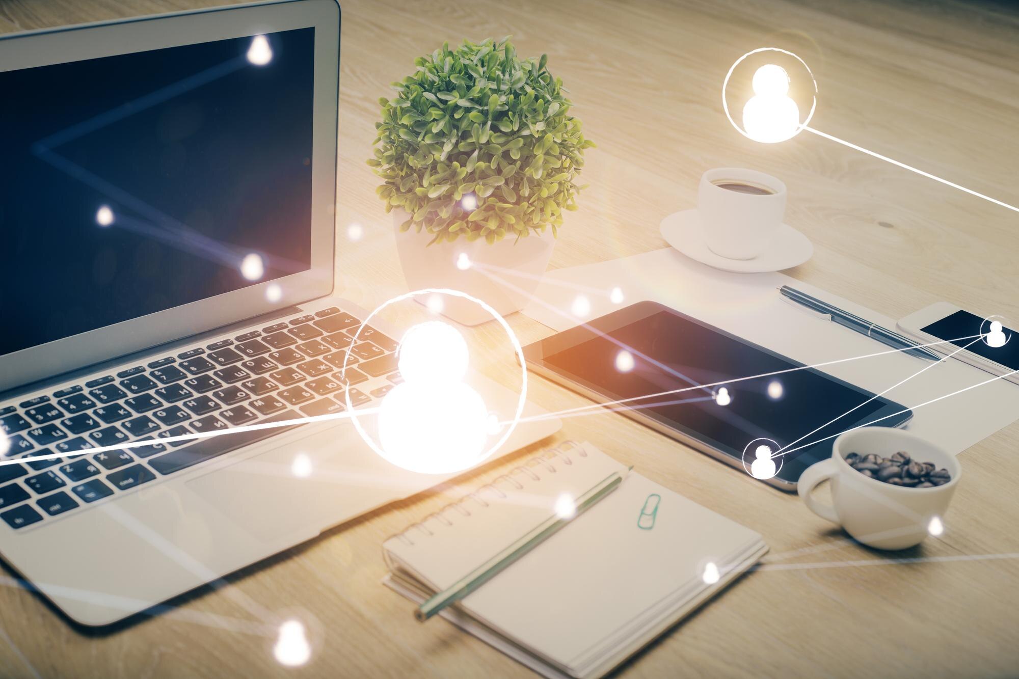 Sosiale nettverk hologram på skrivebord med datamaskin Bilde av en datamaskin med hologram av sosiale nettverk. Laptopen er åpen på et skrivebord med en plante, en kopp kaffe, en notisbok og et nettbrett, omgitt av hologram-lignende sosiale nettverksikoner.