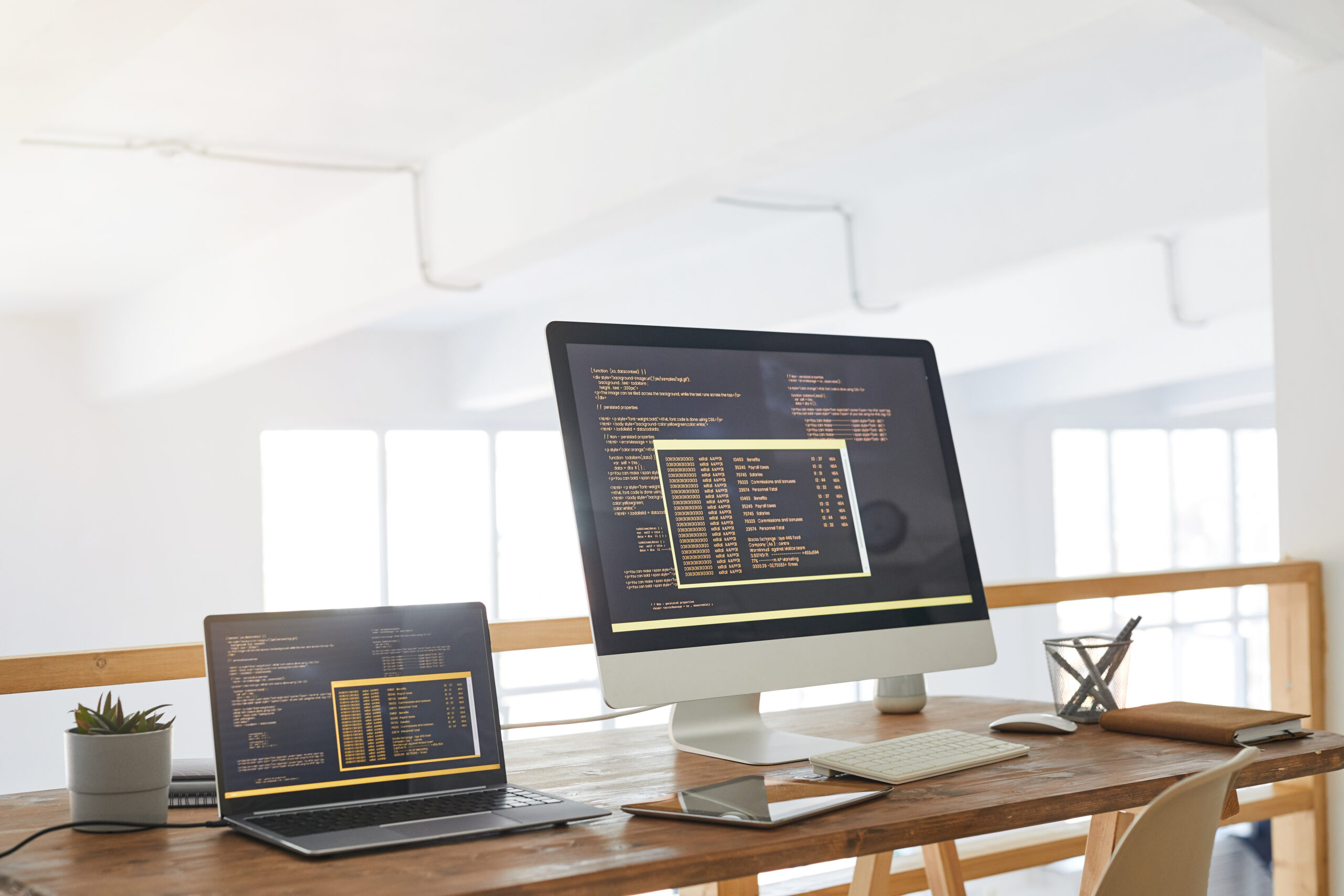Moderne arbeidsplass med en laptop og en stasjonær skjerm, begge viser kode på svart bakgrunn. Naturlig lys strømmer inn.