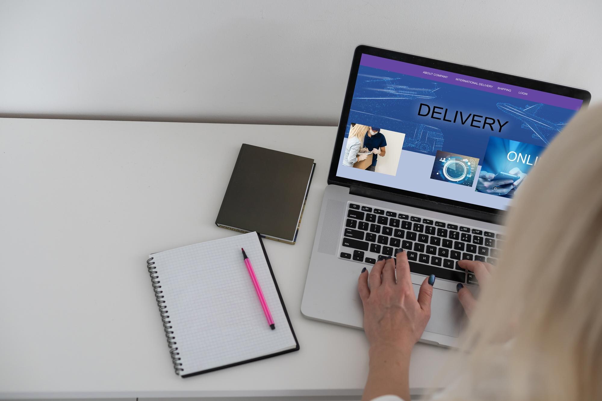 Tittel: Kvinnehender på laptop med leveringsinformasjon Oppsummering: Kvinne jobber på laptop med fokus på leveringsnettsted. Kvinnelige hender på en bærbar datamaskin som viser en nettside med teksten "DELIVERY". Notatblokk og penn ligger ved siden av.